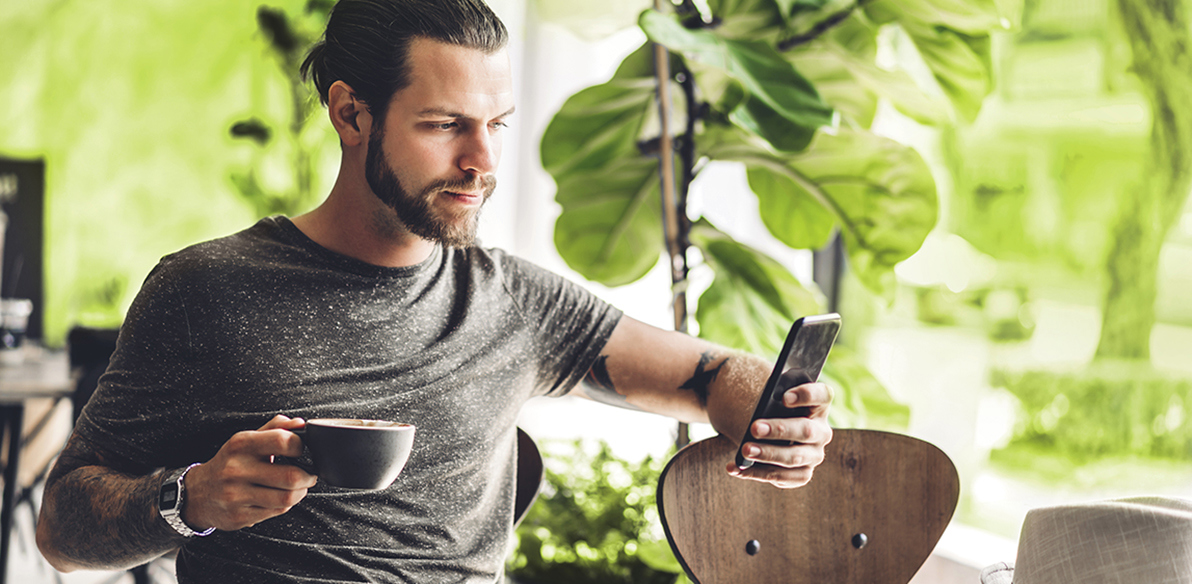 Immagine di un giovane uomo, seduto ad un tavolo: in una mano tiene una tazza di caffé e con l'altra usa il proprio smartphone